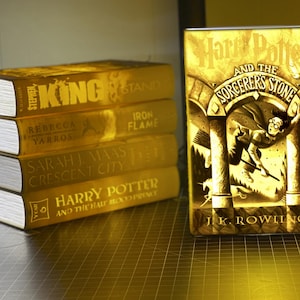 May include: A stack of gold-colored books, including titles by Stephen King, Rebecca Yarros, and Sarah J. Maas. A glowing book cover of "Harry Potter and the Sorcerer's Stone" by J.K. Rowling is also visible.