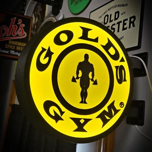 May include: A round, illuminated sign for Gold's Gym. The sign is yellow with black lettering and a silhouette of a weightlifter. The sign is mounted on a wall with other vintage signs in the background.