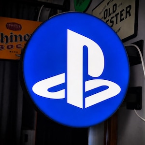 May include: A round, illuminated sign with a blue background and a white PlayStation logo. The sign is mounted on a wall with other vintage signs in the background. A white power cord is plugged into the sign.