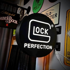 Puede incluir: Un letrero redondo negro e iluminado con la palabra "LOCK" en blanco y en negrita, y la palabra "PERFECTION" debajo. El letrero está montado en una pared, con otros letreros visibles en el fondo.