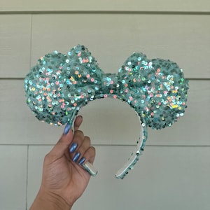 May include: A pair of light blue sequined Minnie Mouse ears with a large bow. The bow is made of iridescent sequins that shimmer in the light.