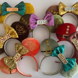 May include: A collection of character-themed ear headbands. Each headband features a large sequined bow with a character name like "Bashful," "Happy," and "Sleepy." The ears are various colors, including red, green, and brown, with gold accents. The headbands are designed for themed events.