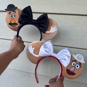 May include: Two novelty headbands featuring Mr. Potato Head designs. One has a blue headband with a black bow, the other has a red headband with a white bow. Both have plush potato-shaped ears with character details.