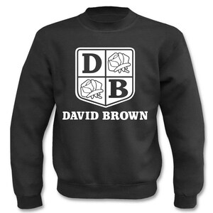 Könnte beinhalten: Schwarzer Sweatshirt mit einem weißen Wappen, das die Buchstaben "DB" und zwei stilisierte Rosen zeigt. Der Text "DAVID BROWN" ist unter dem Wappen gedruckt.