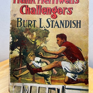 May include: A vintage book cover with the title "Frank Merriwell's Challengers" by Burt L. Standish. The cover features an illustration of two boys jumping over a fence.