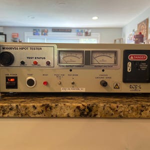 May include: A beige M100BVS5 HIPOT Tester with a black dial, gauges, and warning labels. It has a "TEST STATUS" section with "READY", "TESTING", and "FAIL" indicators. The device includes a "DANGER" label and the text "HIGH VOLTAGE DURING TEST".
