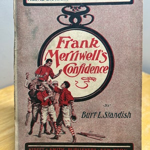May include: A vintage book cover with a red and white illustration of a baseball team. The title is "Frank Merriwell's Confidence" by Burt L. Standish. The book is part of the Medal Library series, number 359.