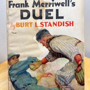 May include: A vintage book cover with an illustration of two baseball players in action. The book title is "Frank Merriwell's Duel" by Burt L. Standish. The book is part of the Merriwell Library series, number 46.