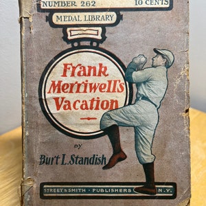 May include: A vintage book cover with a brown background and a circular illustration of a baseball player in a white uniform. The title is "Frank Merriwell's Vacation" by Burt L. Standish. The book is part of the Medal Library series and is numbered 262.