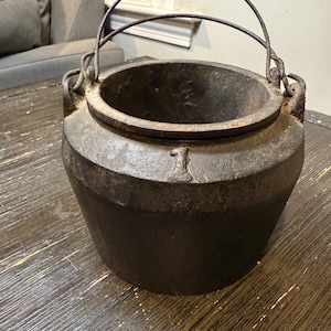May include: A dark, cast iron cauldron with a curved handle and a number "1" etched on the side. The pot has a wide opening and a rounded base. The cauldron is sitting on a dark wooden surface.