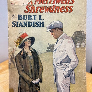 May include: A vintage book cover with an illustration of a man and woman in a golf setting. The title of the book is "Merriwell's Shrewdness" by Burt L. Standish. The book is numbered 65.