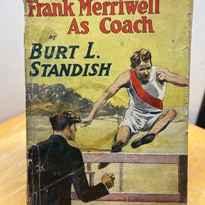 May include: A vintage book cover with a yellow background and a red and white illustration of a man jumping over a hurdle. The book title is "Frank Merriwell As Coach" by Burt L. Standish. The book is part of "The Merriwell Series" No. 72.