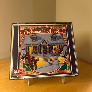 Puede incluir: Un CD de Navidad en América con una ilustración festiva de una casa cubierta de nieve con villancicos cantando frente a ella. El CD es de Reader's Digest y está remasterizado digitalmente.