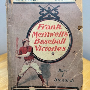 May include: A vintage book cover with a red, white, and blue color scheme. The cover features a baseball player in a red uniform and a white baseball cap. The title of the book is "Frank Merriwell's Baseball Victories" by Burr L. Standish. The book is part of the Medal Library series, number 350.