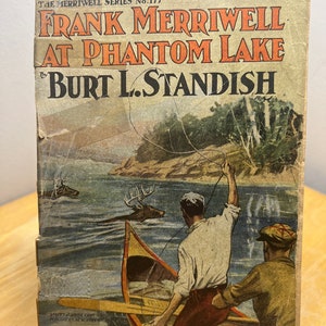 May include: A vintage book cover featuring an illustration of two people in a canoe on a lake. The book title is "Frank Merriwell at Phantom Lake" by Burt L. Standish. The book is part of "The Merriwell Series" number 177.