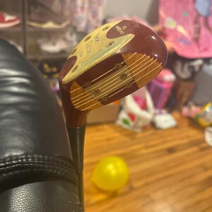 以下が含まれることがあります： 茶色と金色のヘッドを持つ、ビンテージの木製ゴルフクラブ。ヘッドには「The MacGregor Classic」と「3 Wood」の文字が書かれています。