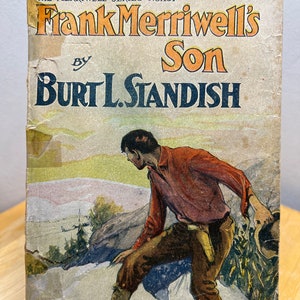 May include: The cover of a vintage paperback book titled "Frank Merriwell's Son" by Burt L. Standish. The book is part of "The Merriwell Series" and is numbered 137. The cover features an illustration of a man in a red shirt and brown pants walking on a rocky path.