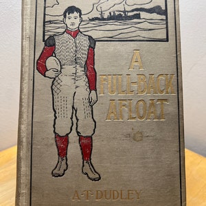 Pode incluir: Uma capa de livro vintage com fundo bege e letras douradas. O título é "A Full-Back Afloat" de A.T. Dudley. A capa apresenta uma ilustração em preto e branco de um jogador de futebol americano segurando uma bola.