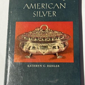 May include: A book titled "AMERICAN SILVER" from "The American Arts Library" series. The cover features a silver serving dish on a red background. The author's name, Kathryn C. Buhler, is printed below the image. The book has a dark teal cover.