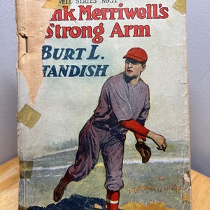 May include: A vintage book cover with a baseball player in a red cap and grey uniform throwing a ball. The title is "Frank Merriwell's Strong Arm" by Burt L. Standish. The book is part of the "Well Series" No. 71.