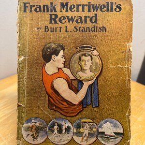 May include: A vintage book cover with a gold background and a picture of a young man holding a medal. The book title is "Frank Merriwell's Reward" by Burt L. Standish. The book is part of the New Medal Library series, number 386.