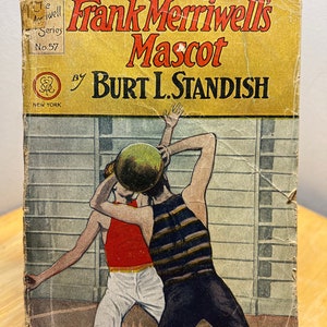 May include: A vintage book cover with a yellow background and the title "Frank Merriwell's Mascot" by Burt L. Standish. The cover features an illustration of two boys playing basketball.