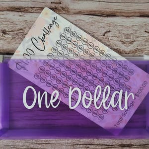 May include: A purple zippered pouch with the words "One Dollar" in white. Inside is a savings challenge card with a floral design and the text "$100 Challenge". The card has circles with the number 69.