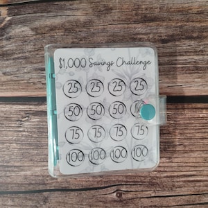 May include: A clear plastic savings challenge binder with a floral design. The binder has a teal button closure and contains a savings tracker with amounts from $25 to $100, totaling $1,000. The text "$1,000 Savings Challenge" is at the top.