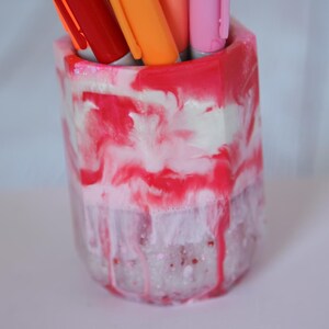 May include: A decorative pen holder with a marbled design in shades of red, pink, and white. The holder contains several pens in red, orange, and pink. The pen holder is a unique desk accessory.