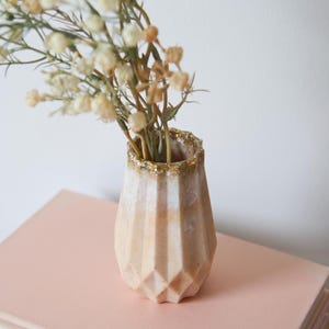 May include: A small, geometric vase in beige and cream, with a gold glitter rim, holds dried flowers. The vase has a faceted design and sits on a pink surface, creating a neutral and elegant aesthetic.