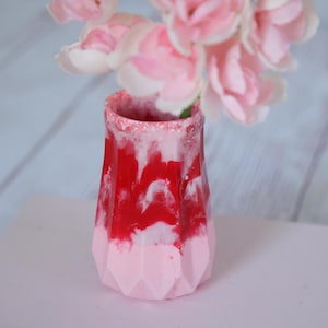 May include: A small, decorative vase with a faceted design. The vase is primarily red with white marbling and a pink base. The rim is speckled with glitter. Pink flowers are arranged inside the vase, creating a floral display.