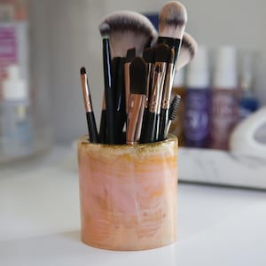 May include: A pink and peach-toned cylindrical makeup brush holder with gold glitter detailing. The holder contains a variety of makeup brushes with black handles and soft bristles, ideal for storing beauty tools.