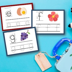 Può includere: Flashcard educative con le lettere e, f e g, ciascuna abbinata a un'immagine di un uovo, un fiore e dell'uva. È visibile anche un portapranzo blu con grafica di mele, una matita e lamponi.