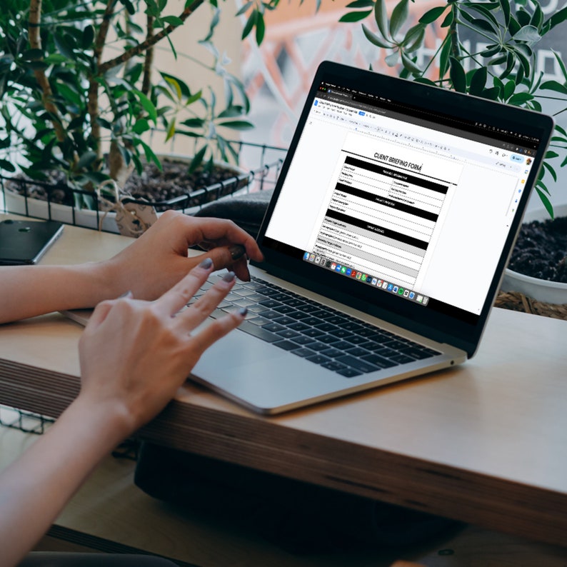 May include: A person working on a laptop computer with a blank "Client Briefing Form" document open on the screen. The laptop is on a wooden table with a potted plant in the background.