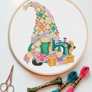 May include: A cross-stitch embroidery project in a wooden hoop featuring a gnome wearing a tall, multi-colored patchwork hat. The gnome is positioned next to a teal sewing machine and a piece of pink and white checkered fabric on a white background.