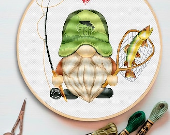 May include: A cross-stitch pattern in a wooden hoop featuring a gnome wearing a green cap with the word FISH and a fish silhouette. The gnome holds a fishing rod with a red lure and a landing net containing a yellow and green fish. Three skeins of green embroidery floss and metal scissors are nearby.