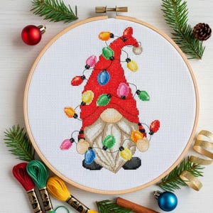 May include: A cross-stitch embroidery in a round wooden hoop featuring a gnome with a tall red hat and a long white beard. The gnome is decorated with colorful Christmas light bulbs in red, blue, green, yellow, and pink, all connected by black lines.
