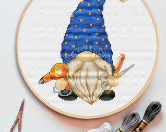 Peut inclure: Un motif de point de croix dans un tambour à broder en bois représentant un gnome à longue barbe, portant un chapeau bleu à pois orange. Le gnome tient un sèche-cheveux orange, un peigne et des ciseaux. Trois échevettes et des ciseaux.