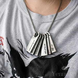 Puede incluir: Un collar de tono plateado con cinco colgantes metálicos rectangulares que cuelgan de una cadena de serpiente. Cada colgante está grabado con distintas ecuaciones matemáticas y fórmulas científicas en tinta negra, creando un aspecto industrial.