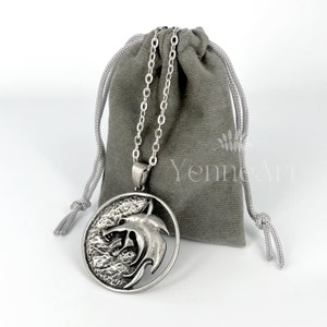 May include: A silver necklace with a round pendant featuring a wolf head design. The pendant is textured and has a detailed wolf head. The necklace comes in a gray velvet pouch with the text "Yenne Art" printed on it.