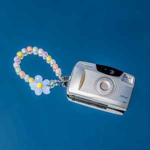 Op de afbeelding: Een zilveren Canon Sure Shot camera met een pastelkleurig kralenbandje en een blauwe bloemenbedel. De camera heeft een zoomlens en staat op een blauwe achtergrond.