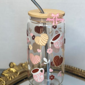 May include: A clear glass water bottle with a bamboo lid and a stainless steel straw. The bottle has a pink and brown heart pattern with a coffee cup design. The bottle is sitting on a gold tray.