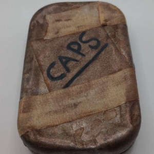 May include: A brown, rectangular container with the word "CAPS" written in black. The container is wrapped with strips of lighter brown material, creating a textured look. The design suggests a vintage or handmade item.
