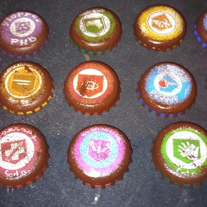 May include: A selection of brown bottle caps, each with a unique design and text in various colours. The caps display symbols and text in red, white, blue, green, and yellow. They are arranged on a dark surface.