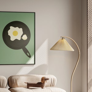 Puede incluir: Una impresi&oacute;n enmarcada con una ilustraci&oacute;n minimalista de un huevo frito en una sart&eacute;n sobre un fondo verde claro. La obra de arte est&aacute; en un marco negro y cuelga sobre un sof&aacute; beige. Un perro salchicha decorativo y una l&aacute;mpara de pie tambi&eacute;n est&aacute;n en la imagen.
