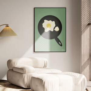 Puede incluir: Un cuadro enmarcado con una ilustraci&oacute;n minimalista de un huevo frito en una sart&eacute;n negra sobre un fondo verde. La obra de arte se exhibe en un sal&oacute;n moderno con un sill&oacute;n beige y una l&aacute;mpara de pie.