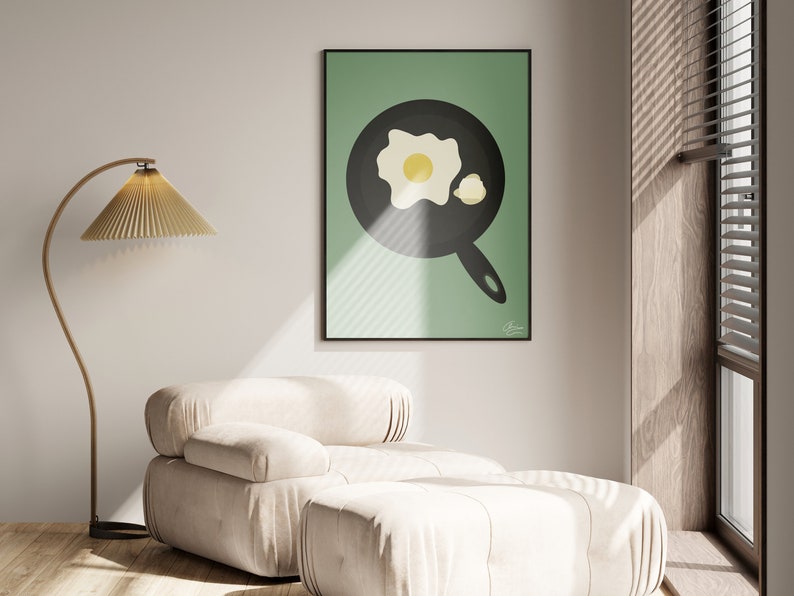 Puede incluir: Un cuadro enmarcado con una ilustraci&oacute;n minimalista de un huevo frito en una sart&eacute;n negra sobre un fondo verde. La obra de arte se exhibe en un sal&oacute;n moderno con un sill&oacute;n beige y una l&aacute;mpara de pie.