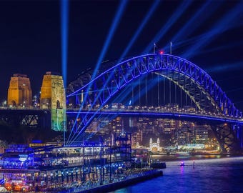 May include: Nighttime view of the Sydney Harbour Bridge illuminated with blue lights. The bridge's arch is lit up, with beams of light radiating upwards. Buildings and city lights are visible in the background.