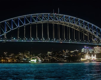 May include: Nighttime view of a large bridge with a blue and white illuminated arch. The bridge spans across a body of water, with city lights and buildings visible in the background. The water is a dark blue color.