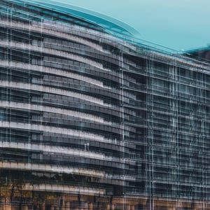 May include: A photograph of a modern building with a curved facade. The building has a glass and steel exterior, with horizontal lines and a teal sky. The image has a blurred effect, creating a sense of movement.